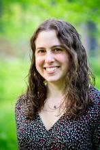 Dr. Gili Freedman, person with long brown curly harir smiling in front of a blurred image of greenery