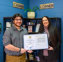 Two people are smiling and holding a framed certificate in a room with blue walls. Shelves with books and plants are in the background. Signs on the wall read "Caring Corner."