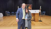 A man in a suit stands next to a woman holding an award on a wooden stage with a religious backdrop featuring a crucifixion image and candles. Empty chairs are arranged behind them.