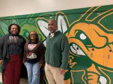 Three people stand smiling in front of green lockers with a large yellow and green bee mascot painted on them. The two women are on the left, one in a graphic T-shirt and blazer, the other in glasses and a cardigan. The man on the right wears a green sweater.