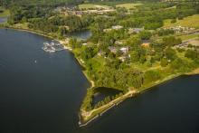 Aerial view of a picturesque landscape featuring a lush, green shoreline surrounded by water. The area includes buildings, roads, and a marina with boats, all encompassed by dense tree cover and grassy fields.