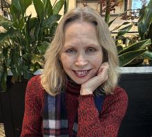 A woman with shoulder-length blonde hair and a red patterned sweater smiles, resting her chin on her hand. 