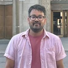 Man wearing glasses and a light pink shirt stands in front of an ornate building entrance.