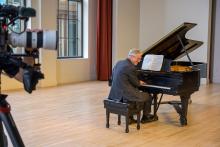 A person wearing a gray suit plays a grand piano in a spacious, well-lit room with large windows and beige walls. A video camera on a tripod is partially visible on the left, recording the performance.