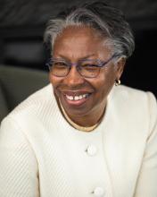 Smiling woman wearing glasses and a white textured blazer, sitting in front of a dark background. Her hair is short and gray, and she is accessorized with small gold hoop earrings.