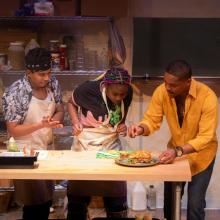 Three people stand at a kitchen counter with food. One person in a yellow shirt points at a dish, while two others wearing aprons watch intently. Shelves with cooking supplies are in the background.