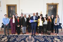 A group of people, dressed in business attire, stand together in a formal room with paintings on the walls. One person in the center holds a St. Mary's sign.
