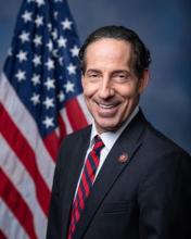 A man in a dark suit and striped tie stands in front of a U.S. flag, smiling at the camera against a blue background.