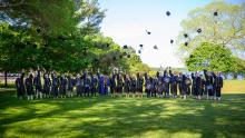 A group of graduates in caps and gowns stand on a lawn, tossing their caps into the air in celebration, surrounded by green trees under a clear sky.