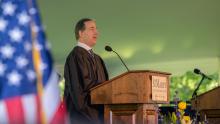 A man in academic robes speaks at a podium labeled "St. Mary’s College" during an outdoor event, with an American flag visible in the foreground.