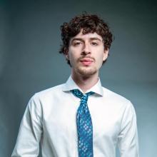 A young man with curly hair wearing a white shirt and blue patterned tie stands against a plain gray background, looking directly at the camera.