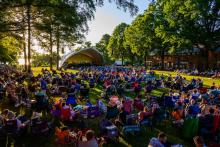 Large crowd sitting on lawn chairs and blankets at an outdoor concert in a park, with a stage set up under trees on a sunny day.