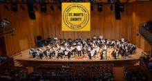 A symphony orchestra performs on stage in a wooden concert hall with a banner overhead reading "Music for Maryland, St. Mary's County, Aug. 1, 2025." Audience members are seated below.