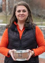 A woman in an orange shirt and black vest stands outdoors, holding a silver bowl with both hands and looking at the camera.