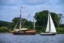 A wooden tall ship with a race committee flag and a modern white sailboat are side by side on a river, with trees and greenery in the background.