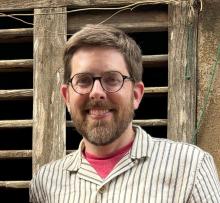 A bearded man with glasses and short hair, wearing a striped shirt and red t-shirt, stands in front of a weathered wooden window.