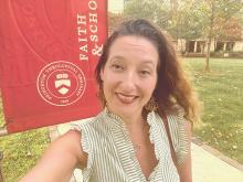 A woman smiles for a selfie in front of a Princeton Theological Seminary banner outdoors on a sunny day.