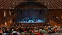 A symphony orchestra performs on stage in a wooden concert hall, with audience members seated on the main floor and balconies.