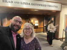 Two people smiling for a selfie inside a theater lobby with a sign reading “Brad and Linda Gottfried Theater” above them; several others are in the background.