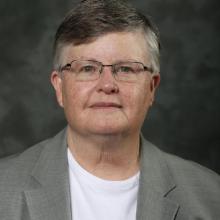 A person with short gray hair, glasses, and a gray blazer over a white shirt poses in front of a dark, neutral background.
