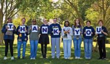 Eight people stand outdoors in a row, each holding a letter sign that together spell out "THANK YOU," with trees and greenery in the background.