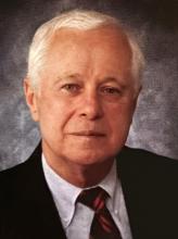 An older man with short white hair, wearing a black suit, white shirt, and red-and-black striped tie, poses against a textured gray background.
