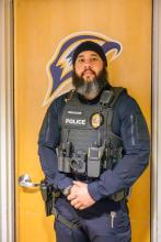 A police officer wearing a tactical vest stands in front of a wooden door with a sports team logo.