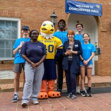 A group of six people and a person in a St. Mary's mascot costume pose in front of Dorchester Hall, smiling for the photo.