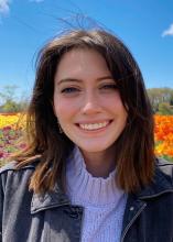 A woman with shoulder-length brown hair smiles outdoors in front of colorful flowers under a blue sky.