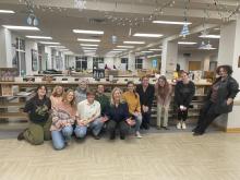 A group of students and a professor pose and smile for a photo in the Hilda C. Landers Library, with artist books and winter-themed decorations visible in the background.