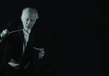 An older man in formal attire holds a conductor’s baton and poses against a dark background, appearing to conduct music.