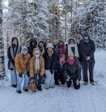 A group of fourteen people dressed in winter clothing pose for a photo on a snowy path surrounded by snow-covered trees.