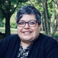 A person with short gray hair and glasses smiles outdoors, wearing a black and white patterned top and a black jacket. Trees and greenery are in the background.
