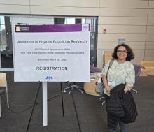 A woman stands next to a registration sign for the "Advances in Physics Education Research" symposium at the University at Albany, dated April 18, 2026.