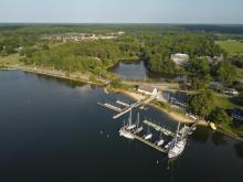 Aerial view of a marina with sailboats docked at piers, surrounded by shoreline buildings, trees, and a pond, with a forested area and fields in the background.
