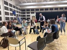 A group of students sits in a circle in a classroom while the instructor (Villada) stands and speaks; another person stands nearby and takes a photo with their phone.