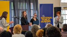 A woman receives an award on stage during a National Public Honors College event, while others applaud and a banner stands in the background.