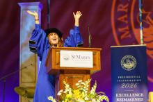 A person in academic regalia stands at a podium with arms raised, speaking at a St. Mary’s College of Maryland event, with a “Presidential Inauguration 2026” sign nearby.