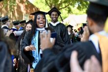 Graduates in caps and gowns walk in a procession outdoors, smiling and celebrating as people applaud around them.