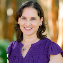 A woman with shoulder-length brown hair, wearing a purple blouse and butterfly necklace, smiles at the camera with a blurred outdoor background.