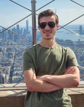 A man in sunglasses and a green shirt stands with arms crossed on an observation deck, with a city skyline and skyscrapers in the background.