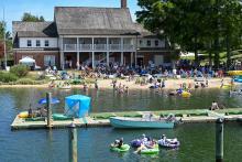 A crowd gathers at a lakeside beach near a large building, with people swimming, lounging on inflatables, and boats docked at a pier in the foreground.