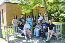 A group of eleven people pose for a photo outside on benches in front of a brick building and trees on a sunny day.