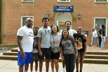 A group of six people stand and smile in front of a brick building with a “Welcome to Caroline Hall” sign above the door.