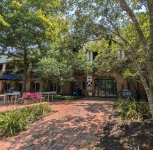 A brick walkway leads to a building entrance surrounded by trees and plants. A sign reads "Campus Center." Tables and pink flowers are visible on the left.
