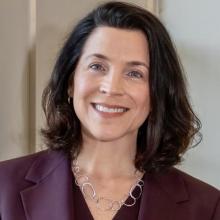 Woman with shoulder-length dark hair wearing a maroon blazer and a silver necklace, smiling in front of a neutral background.