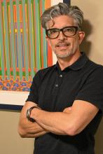 A man with gray hair, glasses, and a black polo shirt stands with arms crossed in front of a colorful abstract painting on a beige wall.