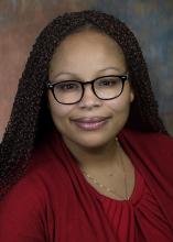 A woman with brown hair and glasses is wearing a red top, smiling at the camera against a neutral background.