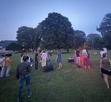 A group of people gathers on a grassy field at dusk, some standing near telescopes, while others form smaller groups. Trees and buildings are in the background.