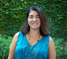 A woman with shoulder-length brown hair, wearing a sleeveless teal top and a green beaded necklace, stands outside in front of a leafy green background.